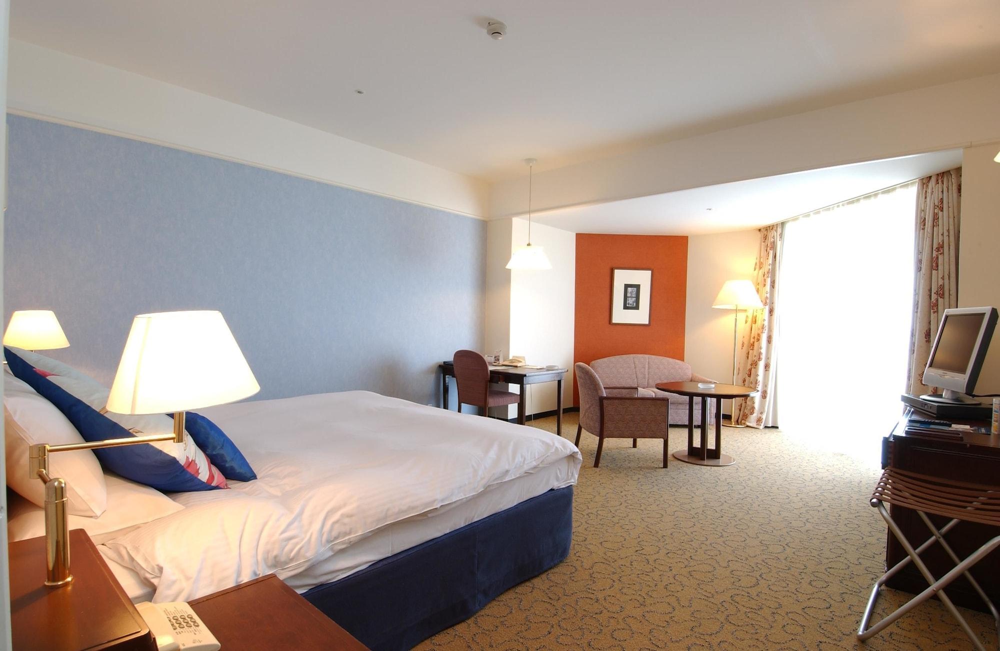 Kobe Portopia Hotel Zimmer foto The photo shows a spacious hotel room with a large bed, featuring white linens and blue accents. There are two bedside lamps providing soft lighting. Next to the bed, there is a small table and a couple of chairs, creating a cozy seating area. A larg