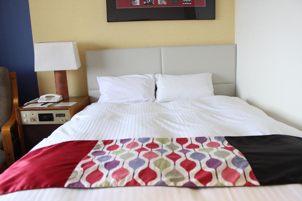 Kobe Portopia Hotel Exterior foto The photo shows a neatly made bed with white bedding and two pillows. At the foot of the bed, there is a decorative blanket featuring a pattern of colorful shapes in various hues. To the side of the bed, there is a bedside table with a lamp, a teleph