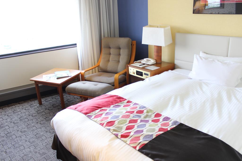 Kobe Portopia Hotel Exterior foto The photo shows a comfortable hotel room. There's a bed with a white comforter and a decorative blanket featuring geometric patterns at the foot. To the side, there’s a small table with a lamp and a phone. Next to it is a cozy armchair. A window lets
