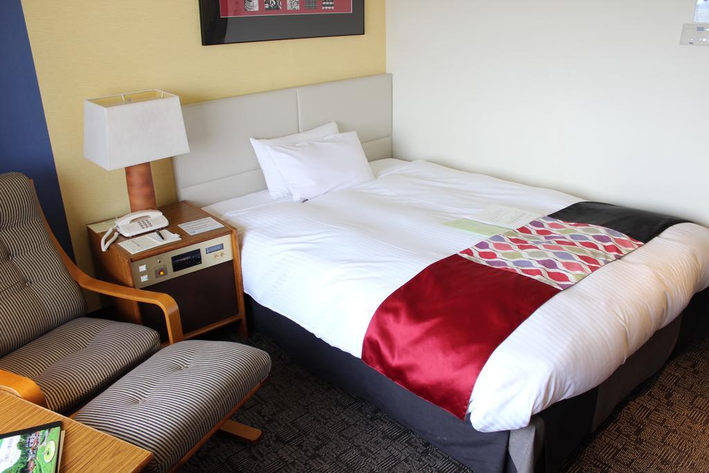 Kobe Portopia Hotel Exterior foto The photo shows a small hotel room featuring a neatly made bed with a colorful throw across the bottom. Next to the bed, there is a nightstand with a lamp and a phone. In the corner, there's a comfortable armchair positioned beside a wooden side tabl