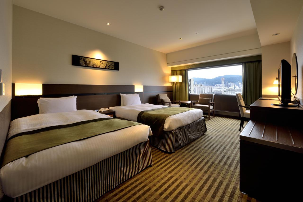 Kobe Portopia Hotel Exterior foto The photo shows an interior of a hotel room. There are two beds with white linens and dark green throws. A large window offers a view of the outside, and there is a small table and chair positioned nearby. The room features warm lighting, wooden furn