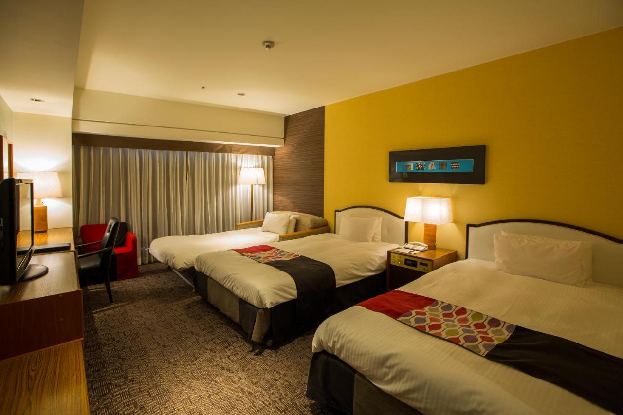 Kobe Portopia Hotel Exterior foto The photo shows a hotel room featuring three beds arranged in two separate areas. Each bed has a combination of white bedding and colorful accents. There are bedside lamps providing soft lighting on either side of the beds. A large window, dressed wi